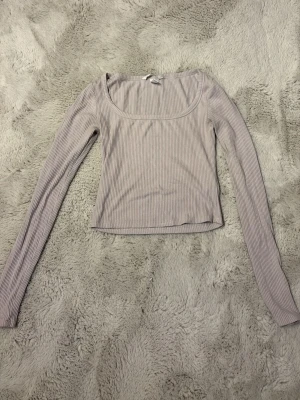 Ljusgrå ribbad långärmad topp - Säljer en ljusgrå ribbad topp med lång ärm och rundad, djup urringning. Toppen är croppad och har en slim passform som sitter snyggt på kroppen. Perfekt att styla med jeans eller kjol för en clean look.
