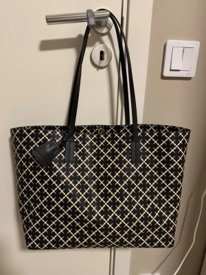 Svart och beige shoppingväska Marlene Birger - Snygg shoppingväska från By Malene Birger i svart och beige med rutigt mönster. Väskan har långa svarta handtag och en tag med logga. Tillverkad i tåligt syntetmaterial och har en rymlig form som rymmer allt du behöver.