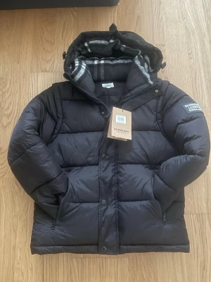 Svart pufferjacka från Burberry - Säljer en svart pufferjacka från Burberry med klassiskt rutigt foder i huvan. Jackan har dragkedja, knappar och två fickor framtill. Stort Burberry-tryck på ärmen och en riktigt clean look. Perfekt för kalla dagar och riktigt snygg till streetwear.
