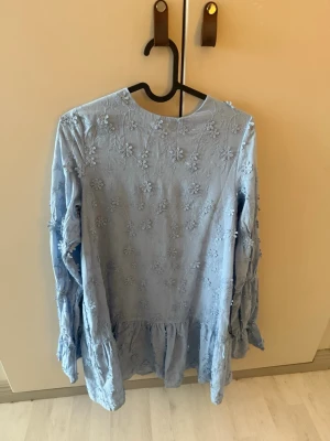 Ljusblå blus med broderade blommor - Supersöt ljusblå blus med broderade blommor och volang nertill. Blusen har långärmade ärmar med rynkade detaljer och en djup v-ringning med knytband framtill. Perfekt för en trendig och romantisk look.bara testad aldrig använd