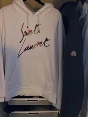 Vit Saint Laurent hoodie med tryck - Säljer en vit hoodie från Saint Laurent med färgglatt logotyp-tryck på bröstet. Hoodien har huva med dragsko och en stor magficka framtill. Perfekt för dig som vill ha en stilren men ändå unik look. Materialet känns mjukt och bekvämt.