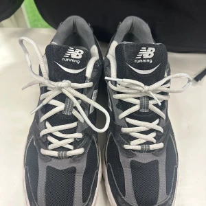 Svarta New Balance sneakers 45 - Snygga svarta och grå New Balance sneakers med vit logga på sidan och tungan. Skorna har vit snörning och en platt sula. Ovandelen är i mesh och syntetmaterial för en sportig look. Perfekta för dig som gillar streetwear och vill ha bekväma skor till vardags.