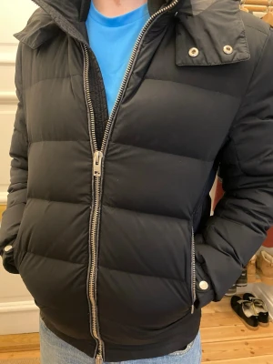 J.Lindeberg vinterjacka - Snygg svart pufferjacka med avtagbar huva och dubbla dragkedjor framtill. Jackan har quiltad design, långa ärmar och ribbade muddar. Perfekt för kalla dagar och ger en clean look till din outfit. Storlek S passar S.KOM MED PRISFÖRSLAG