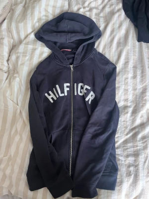 Marinblå Hilfiger hoodie med dragkedja - Snygg marinblå hoodie från Tommy Hilfiger med vit logga över bröstet. Hoodien har dragkedja framtill, två fickor och en klassisk huva. Tillverkad i mjukt bomullsmaterial som är skönt att ha på sig. Perfekt för en avslappnad och trendig look.