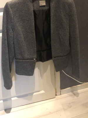 Grå kavaj från Vero Moda med dragkedja - Snygg grå kavaj från Vero Moda med raka linjer och dragkedjedetaljer framtill. Kavajen har lång ärm och är fodrad med svart tyg på insidan. Perfekt för en stilren och modern look.