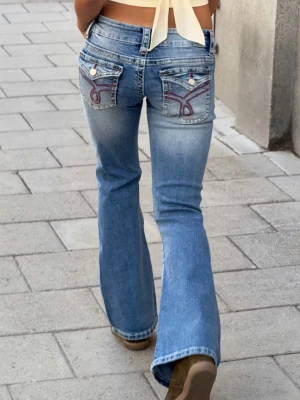 Blå bootcut jeans med broderade fickor - Säljer ett par ljusblå bootcut jeans med snygga broderade detaljer på bakfickorna och låg midja.Perfekta för en trendig och avslappnad stil💗