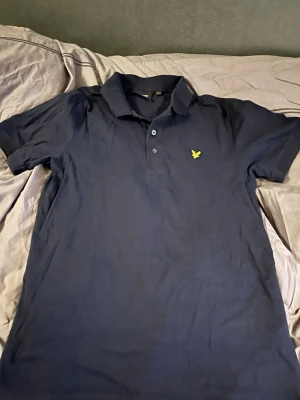 Marinblå pikétröja från Lyle & Scott - Snygg marinblå pikétröja från Lyle & Scott med klassisk krage och knappar framtill. Tröjan har det ikoniska gula logobroderiet på bröstet och är tillverkad i mjuk bomull. Perfekt för en stilren och avslappnad look.