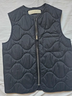 Svart quiltad väst från ARKET - Stilren svart quiltad väst från ARKET med vågigt mönster, dragkedja framtill och en bröstficka. Västen har en modern, boxig passform och är tillverkad i ett lätt, vadderat material som passar perfekt för lager-på-lager-stil. Ingen prislapp kvar, men aldrig använd, alltså helt ny. 