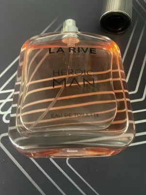 La Rive Heroic Man Eau de Toilette - La Rive Heroic Man Eau de Toilette är en parfym för män i en stilren, transparent glasflaska med svarta detaljer och text. Doften har en fräsch och maskulin känsla, perfekt för dig som vill sticka ut. Flaskan har en metallfärgad kork och innehållet har en lätt orange ton.