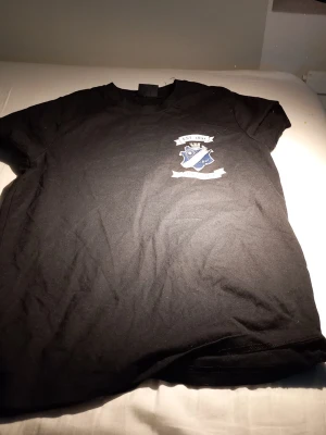 Svart AIK t-shirt med tryck - Svart t-shirt från AIK med klubbens klassiska logga och texten 'EST 1891' samt 'STOCKHOLM' tryckt på bröstet. T-shirten har rund hals och korta ärmar, perfekt för dig som vill visa ditt AIK-stöd. Med storlek Small.