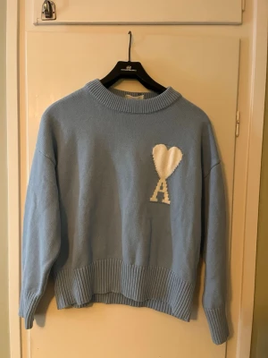 Ljusblå stickad ami paris tröja - Ljusblå stickad Ami paris tröja med ribbade muddar och nederkant. På bröstet finns en vit hjärtdetalj och bokstaven 'A' i samma färg. Tröjan har avslappnad passform och rund halsringning, perfekt för flesta stilar!