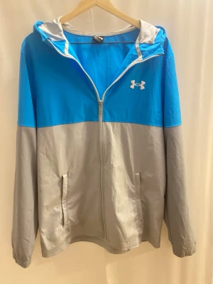 Blå och grå vindjacka från Under Armour - Snygg tvåfärgad vindjacka från Under Armour i blått och grått. Jackan har huva, dragkedja framtill och två fickor. Perfekt för blåsiga dagar och enkel att matcha med olika outfits. Tillverkad i lätt polyester för skön känsla.       