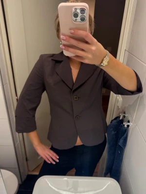 Mörkbrun kavaj med trekvartsärm - Stilren mörkbrun kavaj med trekvartsärmar och två knappar framtill. Klassisk krage och figurnära passform som ger en snygg siluett. Perfekt att styla till både jeans och kostymbyxor.