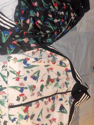 Adidas blommig vindjacka svart/vit - Säljer en snygg vindjacka från Adidas med blommigt mönster i färger som röd, blå, grön och rosa. Jackan har klassiska vita ränder på ärmarna, dragkedja framtill och finns i både svart och vit variant. Perfekt för dig som vill sticka ut med sportig stil. Man kan köpa båda för 1000 eller separat för 600, köptes för 1000 kr styck 