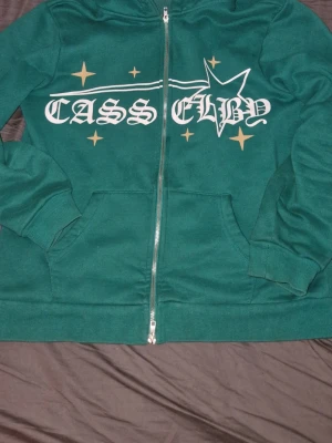 Grön zip hoodie  - Snygg grön hoodie med dragkedja. Tryck i vitt och guld med text och stjärndetaljer på bröstet. Utrustad med huva och två stora fickor framtill. Tillverkad i mjukt och bekvämt material, perfekt för en avslappnad stil.