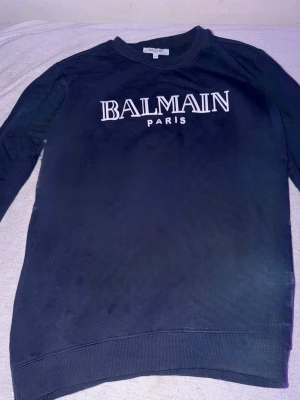Svart sweatshirt från Balmain Paris - Cool svart sweatshirt från Balmain Paris med vit logga tryckt på bröstet. Tröjan har rund halsringning, långa ärmar och ribbade muddar vid ärmslut och nederkant. Perfekt för en avslappnad och trendig look.