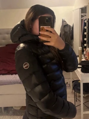 Svart colmar jacka med grå insida  - Snygg svart dunjacka från Canada Snow med hög krage och huva. Jackan har en quiltad design, dragkedja framtill och en patch med logga på ärmen. Perfekt för kalla dagar och ger en cool, sportig look.