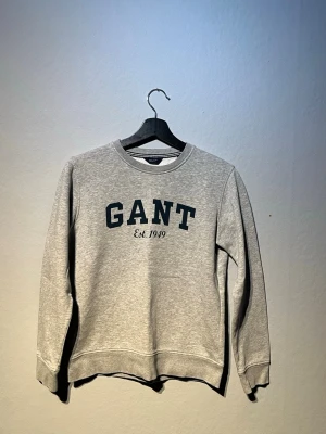  GANT tröja st 164 - Klassisk grå sweatshirt från GANT STORLEK 164med rund halsringning och tryckt logga samt 'Est. 1949' på bröstet. Tröjan har ribbade muddar vid ärmslut och nederkant. Perfekt för en avslappnad och stilren look.