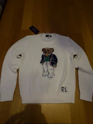 Vit stickad tröja Polo Ralph Lauren - Vit stickad tröja från Polo Ralph Lauren med ikonisk björn på bröstet. Tröjan har rund halsringning, ribbade muddar och RL-broderi nedtill. Perfekt för dig som gillar klassisk och lekfull stil.