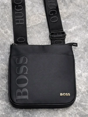 Svart axelväska från Hugo Boss - Snygg svart axelväska från Hugo Boss med justerbar axelrem där det står 'HUGO'. Väskan har en stor BOSS-logga i relief på framsidan och en mindre i guld. Stängs med dragkedja och har en stilren, fyrkantig form.