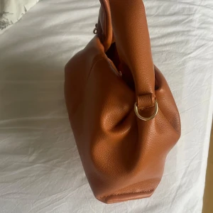 Brun handväska  - Snygg brun handväska i skinn med en stilren design. Väskan har en praktisk storlek och är perfekt för att bära med sig det viktigaste. Den har en enkel och elegant look med rundade handtag.