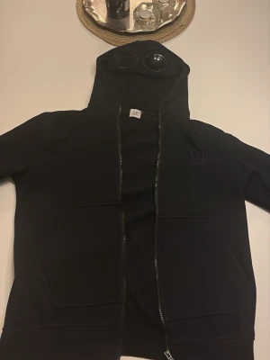 Svart hoodie med glasögon från C.P. Company - Säljer en svart hoodie från C.P. Company med ikoniska inbyggda glasögon i huvan. Hoodien har dragkedja framtill, två stora fickor och ribbade muddar. Tillverkad i mjukt bomullsmaterial och har en clean, modern look med diskret logga på bröstet.