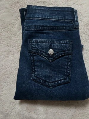 Mörkblå bootcut jeans med fickdetalj - Snygga mörkblå jeans med bootcut passform och cool ficka med knappdetalj bak. Klassisk femficksmodell och bälteshällor. Jeansen har en lätt tvättad look och är tillverkade i mjuk bomullsdenim som sitter skönt hela dagen. Storlek 170 massa som 34/xs