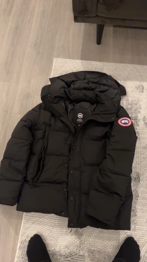 Svart  jacka Canada Goose Wyndham - Säljer en svart jacka  från Canada Goose med röd label.  stor huva med avtagbar pälskant. Jackan har dragkedja och knappar framtill, samt flera praktiska fickor. Perfekt för kalla vinterdagar.