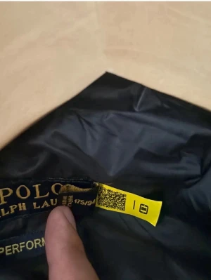 Svart dunjacka från Polo Ralph Lauren - Snygg svart dunjacka från Polo Ralph Lauren med glansig finish och huva. Jackan har klassisk pufferstil med breda, quiltade sektioner och dragkedja framtill. Perfekt för kalla dagar och enkel att matcha med olika outfits.