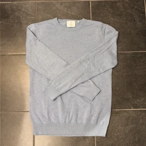 Ljusblå tröja från Bläck, storlek S - En stilren ljusblå tröja från Bläck i storlek S. Tröjan har rund halsringning, långa ärmar och ribbade muddar vid ärmslut och nederkant. Perfekt för lager-på-lager och enkel att matcha med jeans eller chinos.