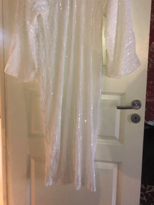 Vit glittrig långklänning - Säljer en vit långklänning med glittriga detaljer och vida ärmar. Klänningen har en v-ringad hals och ett elegant fall. Perfekt för dig som vill sticka ut med en glamorös look.