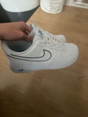 Nike Air Force 1 vita sneakers - Nästan aldrig använda äkta airforce köpta i barcelona Nike affär Säljer ett par klassiska Nike Air Force 1 sneakers i vitt skinn med blå detaljer och svart swoosh. Skorna har snörning, perforerad tå och låg profil. Perfekta för dig som vill ha en clean och tidlös look.