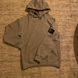 Grå hoodie från Stone Island - Grå hoodie från Stone Island med klassisk huva och snörning. Tröjan har ribbade muddar och en svart patch med logga på vänster ärm. Perfekt för en avslappnad och stilren look. Den är liten för mig och jag är 187cm 