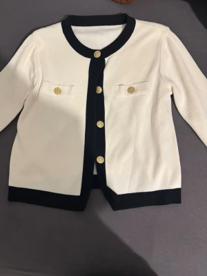 Vit kofta med svarta detaljer - Stilren vit kofta med svarta kanter och guldknappar framtill. Två dekorativa fickor med guldknapp på bröstet. Klassisk och tidlös design, perfekt för att lyfta vilken outfit som helst.