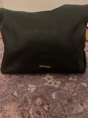 Svart handväska från Calvin Klein - Stilren svart handväska från Calvin Klein i mjukt skinn. Väskan har minimalistisk design med diskret logga i silver framtill. Perfekt för dig som gillar klassiska och enkla accessoarer. Säljer eftersom den ej kommer tillanvönding. Först var det en annan kedja på väskan men handtaget är från en annan väska. Den funkar bra och som ni ser är den fast med ett litet hårband haha. De går att ta av la den där för då är den ännu bättre. Köpte den för 1 år sen. 