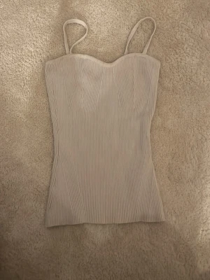 Beige ribbad linnetopp med smala band - Snygg beige ribbad linnetopp med smala axelband och figurnära passform. Toppen har en lätt hjärtformad urringning och är tillverkad i ett stretchigt material som sitter bekvämt. Perfekt att styla med jeans eller kjol.