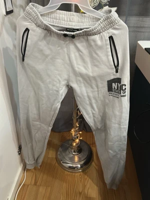 Grå mjukisbyxor från Reaction NYC - Snygga grå mjukisbyxor från Reaction med tryck 'NYC State of Mind' på ena benet. Byxorna har svarta dragkedjor på fickorna och elastisk midja med snörning. Perfekta för en chill och avslappnad stil.