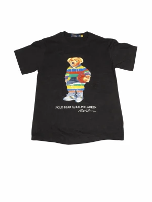 Svart Polo Bear t-shirt Ralph Lauren - Svart t-shirt från Ralph Lauren med ikoniska Polo Bear-trycket framtill. Björnen har färgglada kläder och håller en basketboll. Klassisk rund hals och normal passform. Perfekt för dig som vill ha en stilren men lekfull look. Org pris 1499 mitt pris 599