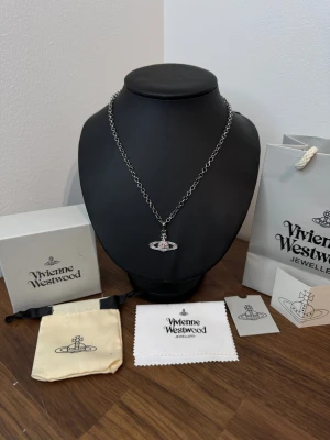 Vivienne Westwood halsband med rosa stenar - Snyggt halsband från Vivienne Westwood med ikonisk orb-berlock dekorerad med rosa glittrande stenar. Kedjan är i silverfärgad metall med rundade länkar och har en graverad logga vid låset. Perfekt statement-piece för dig som gillar coola detaljer och designeraccessoarer.Kommer med: gift bag, gift box, dust cloth, care instructions, dust bag (som på bilden).