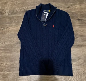 Marinblå stickad tröja Polo Ralph Lauren - Snygg marinblå stickad tröja från Polo Ralph Lauren med klassisk kabelstickning och halv dragkedja vid kragen. Tröjan har ribbade muddar och en röd broderad logga på bröstet. Perfekt för dig som gillar stilren och tidlös design.