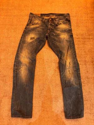 Replay jeans med slitningar och tvättad look - Säljer ett par Replay jeans med coola slitningar och tvättad effekt. Modellen har klassisk femficksdesign, raka ben och snygga detaljer på bakfickorna. Jeansen har en mörkblå ton med ljusare partier och är tillverkade i robust denim. Storlek på Jeansen är 32/34!