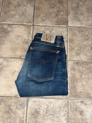 Nudie Jeans - Säljer ett par Nudie Jeans Skinny Lin i mörkblå tvätt med klassiska orange sömmar. Modellen är skinny fit med normal midja och tillverkad i stretchig denim. Snygga detaljer och ikonisk Nudie-logga på bakfickan. Midja 31cm, längd 94cm. JAG SKICKAR EJ FLER BILDER NÄR JAG BÄR JEANSEN! för storleksguide hänvisar jag till måtten och för bättre inblick i passformen rekommenderar jag att googla på modellnamnet. kolla måtten noggrant :) s4 