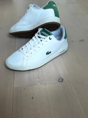 Vita sneakers från Lacoste med gröna detaljer - Snygga vita sneakers från Lacoste med gröna detaljer på häl och plös. Klassisk design med snörning, rund tå och platt sula. Skorna har Lacoste-logga på sidan och är tillverkade i skinn med inslag av mocka på hälen.
