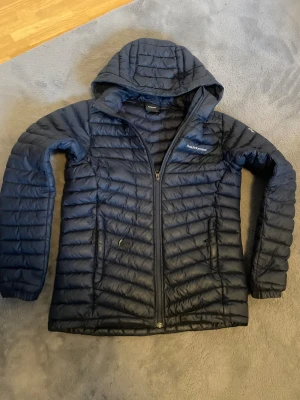 Mörkblå dunjacka Peak Performance - Snygg mörkblå dunjacka från Peak Performance med huva och hel dragkedja. Jackan har quiltad design, två sidofickor med dragkedja och logga på bröstet. Perfekt för kalla dagar och har en clean, sportig look som funkar till streetwear.170