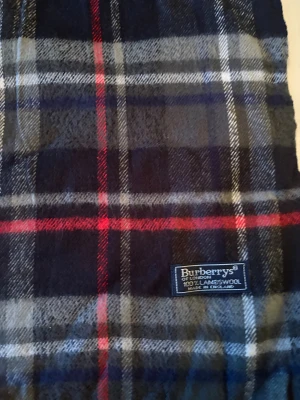 Rutig halsduk från Burberrys i ull - Snygg rutig halsduk från Burberrys i 100% lammull. Färgerna är mörkblå, grå, vit och röd med klassiskt rutmönster. Halsduken har fransar i kanterna och är mjuk och varm, perfekt för kyliga dagar. Märke och material syns tydligt på etiketten.