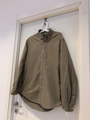 Olivgrön skjorta från Polo Ralph Lauren - Olivgrön skjorta från Polo Ralph Lauren med klassisk button-down krage och orange broderad logga på bröstet. Custom fit-modell med långa ärmar och vita knappar framtill. Skjortan är tillverkad i mjuk bomull och har en stilren, tidlös look.