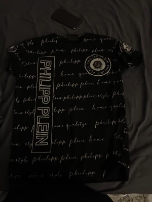 Svart Philipp Plein t-shirt med tryck - Snygg svart t-shirt från Philipp Plein med Platinum Cut. T-shirten har vit textprint över hela plagget och ett stort vertikalt PHILIPP PLEIN-tryck framtill. Rund hals och korta ärmar, samt logomärke på bröstet och ärmen. Perfekt för dig som gillar statement-plagg.