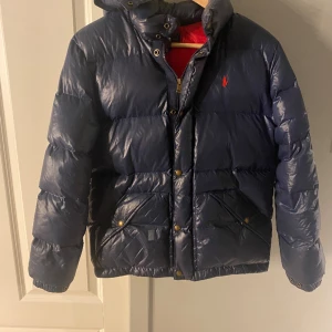 Mörkblå dunjacka från Polo Ralph Lauren - Snygg mörkblå dunjacka från Polo Ralph Lauren med röd insida och klassisk logga på bröstet. Jackan har huva, dragkedja och tryckknappar, samt två stora fickor framtill. Perfekt för kalla dagar och har en riktigt puffig look. Jackan heter glosst down jacket. Det är junior storlek L men sitter ungefär som XXS eller XS jag är 168 cm lång och den blev lite kort men den passar utmärkt från 155cm till 165 cm. Nypris är 3500 men jag säljer min för 2500 är även väldigt öppen till byte  fjällräven jacka