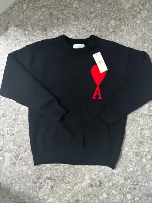 Svart Ami tröja med rött hjärta - Svart tröja från Ami med ett stort rött hjärta och bokstaven A broderat på bröstet. Rund hals och ribbade muddar vid ärmslut och nederkant. Tillverkad i ekologisk bomull enligt GOTS-standard. Perfekt för dig som gillar stilrena detaljer och snygga loggor, passar som en stor M eller en liten L i herrstorlek.