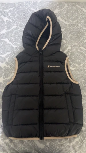 Svart dunväst från Champion med huva - Svart dunväst från Champion med beige kantdetaljer och huva. Västen har quiltad design och dragkedja framtill. Tillverkad i 100% polyester, perfekt för lager-på-lager och chill stil. Champion-logga broderad på bröstet.